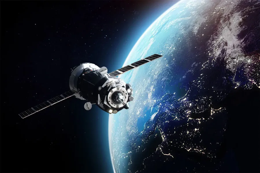 A satellite or spacecraft orbits above Earth, with the planet's illuminated surface and city lights visible below against the darkness of space.